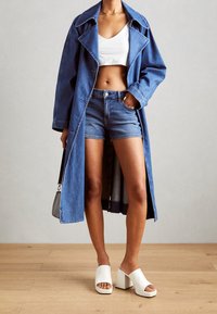 Osoba v modrém denimu trenčkotu, denime šortkách, bílém crop topu a bílých podpatkových mule botách, stojící na dřevěné podlaze.