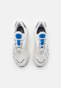 White mesh athletic shoes with gray accents. Features blue branding on the tongue, lace closure, ribbed texture, and a rounded sole.