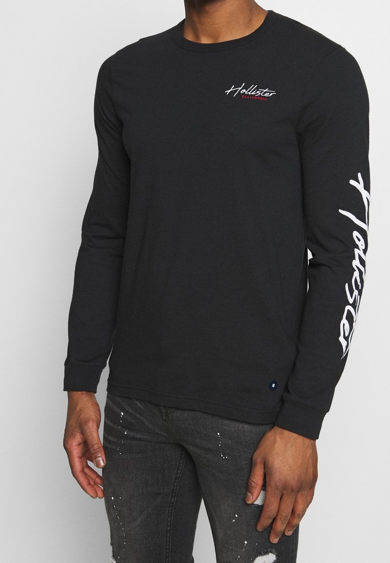 Svart långärmad skjorta i bomull, med en vit grafisk logotyp på ärmen och en mindre broderad logotyp på bröstet.