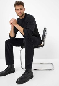 Zwarte leren laarzen, goed passende zwarte corduroybroek en een donkergrijze trui. Het model zit op een stoel met een chroomframe en een houten zitting.