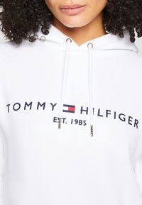 Hvid hættetrøje med "TOMMY HILFIGER"-logo i marineblå med røde og hvide accenter. Guldhardware på snørebåndene tilføjer detaljer.