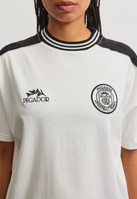 Άτομο που φοράει λευκό T-shirt Pegador με μαύρο ριγωτό γιακά και πάνελ στους ώμους, καθώς και λογότυπα της ομάδας ποδοσφαίρου Pegador στο στήθος.