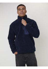 Jonge man in een marineblauwe fleecejas, met de voorkant half dichtgeritst en met één hand in een zak, staand tegen een effen lichte achtergrond.