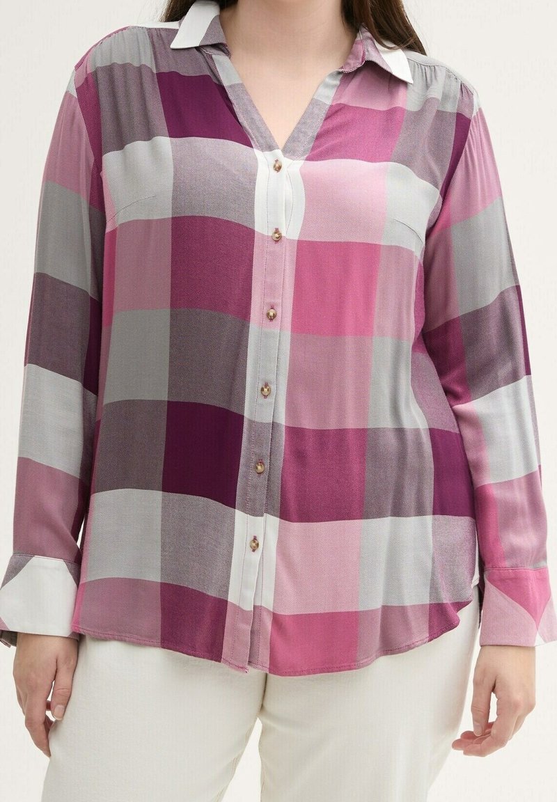 Kvinde iført en langærmet skjorte med knapper, dækket af store pink, lilla og grå tern, sammen med hvide bukser.