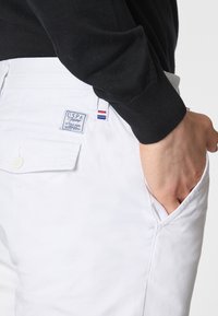 Pantaloni di cotone bianchi con tasche laterali, dotati di un'etichetta USPA visibile sul retro e una manica di un maglione di maglia nera nelle vicinanze.