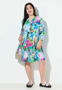 Vestido estampado floral colorido con mangas largas, escote en forma de V y falda de volantes. Combinado con sandalias plateadas y un bolso de hombro metálico.