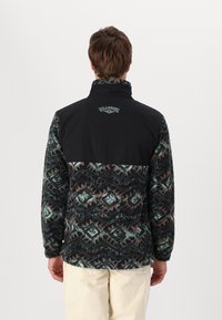 Fleecejack met een zwarte bovenkant en een gepatrooned onderkant in tinten groen, blauw en rood, met een hoge kraag en borduurlogo.