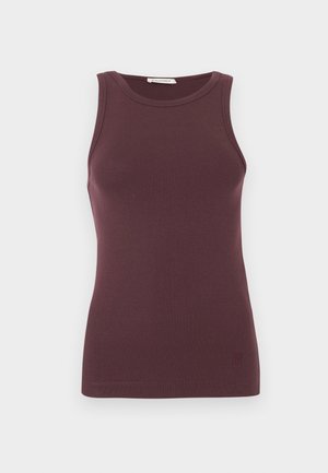 Vrouw met steil zwart haar, bril, draagt een mouwloze bruine top, lichtgroene wijde jeans en witte puntige schoenen, staat met handen in de zij.