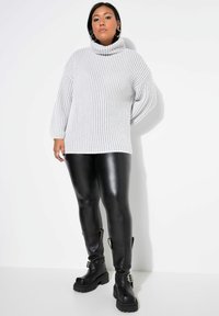 Grauer gerippter Pullover mit hohem Kragen, kombiniert mit schwarzen glänzenden Leggings und klobigen schwarzen Stiefeln mit Schnallen und dicken Sohlen.