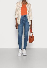 Camisa de patrón beige claro sobre una blusa naranja de cuello en V, combinada con jeans ajustados azules y zapatillas blancas. Lleva un pequeño bolso marrón.