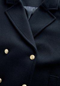 Sötétkék gyapjú kabát dupla gombos fazonnal, arany gombokkal. Kidolgozott hajtókák és sima bélés látható. Texturált anyag közeli képe.