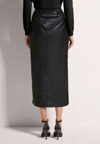 Schwarzer Leder-Midirock mit tailliertem Bund und gerader Silhouette, ausgestattet mit einem glänzenden Finish und seitlichem Schlitz. Kombiniert mit schwarzen High Heels.