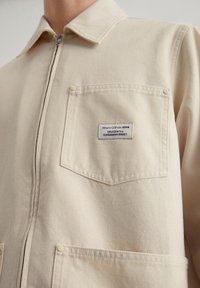 Beige denimjack met een kraag, ritssluiting aan de voorkant, twee zakken en een label met de tekst "Marc O'Polo Denim" op de borstzak genaaid.