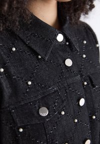 Camicia di denim nera con colletto strutturato, dettagli in metallo argentato decorativo e accenti di perle con motivi geometrici ricamati.