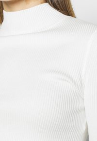 Chemise à col roulé blanche et côtelée mettant en avant un col haut et un tissu texturé. Gros plan sur la partie supérieure du corps, soulignant l'élasticité du matériau.