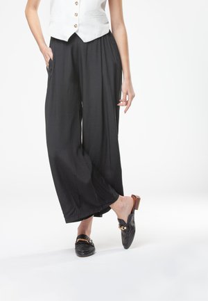 Donna che indossa pantaloni neri a gamba larga, top bianco senza maniche con bottoni e mocassini neri slip-on con dettaglio catena dorata.