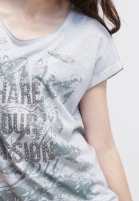 Camiseta de algodón gris claro con mangas cortas, que presenta un estampado gráfico en verde y tachuelas plateadas, y un corte relajado.