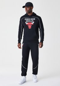 Crna hulahop majica s crvenim logom Chicago Bullsa, bijelim akcentnim crtama, uparena s crnim trenirkama. Bijele tenisice upotpunjuju sportski izgled.