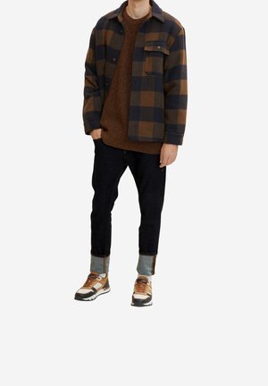 Kariertes Jacket in Braun und Marineblau, Strickpullover, dunkle Jeans mit Aufschlägen und Sneaker mit braunen Akzenten und weißen Details.