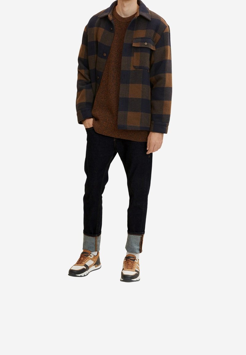Giacca a quadri in marrone e blu, maglione in maglia, jeans in denim scuro con risvolti e sneakers con accenti marroni e dettagli bianchi.