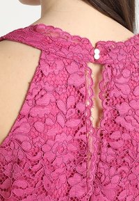 Fuchsia spetsblus med blommönster och vågiga kanter, med knäppning bak och en låg, öppen design.