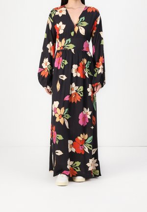 Robe longue noire à fleurs avec des fleurs rouges, roses et jaunes vives. Présente un décolleté en V profond et des manches bouffantes. Portée avec des baskets blanches.