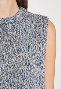 Dzianinowy top bez rękawów w kolorze niebieskim i beżowym, z teksturowanym wzorem i okrągłym dekoltem. Materiał wydaje się miękki i oddychający.