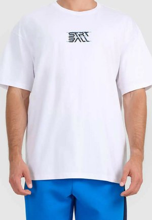 Mężczyzna ubrany w białą koszulkę z małym napisem "START BALL" na piersi oraz jasnoniebieskie spodenki na jednolitym tle.