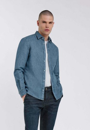 Camicia di denim azzurro chiaro, maniche lunghe, colletto a pulsante, con leggere sbiaditure e dettagli di cucitura. Indossata sopra una maglietta bianca e jeans scuri.