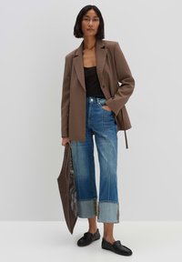 Blazer marrone su una canotta nera, abbinato a jeans blu a gamba larga con orli grezzi e cuciture a contrasto, e scarpe slip-on nere.