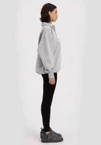 Szary, oversize'owy sweter z kołnierzem i detalami guzików, w połączeniu z czarnymi legginsami i szarymi trampkami z wysoką cholewką, pokazany z profilu.