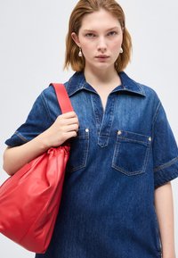 Jeune femme aux cheveux courts portant une chemise en denim bleu et tenant un grand sac à bandoulière en cuir rouge sur un fond uni.