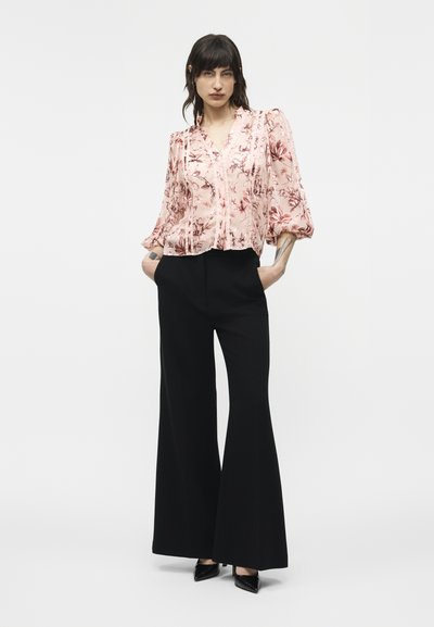 Femme aux cheveux foncés portant un chemisier rose à fleurs avec des manches bouffantes et un pantalon large noir, debout les mains dans les poches.