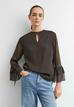 Donna che indossa una camicia marrone scuro trasparente con maniche svasate arricciate e jeans azzurri, in piedi con una mano in tasca contro una parete bianca.