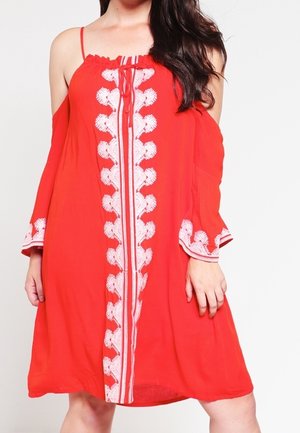 Kvinde iført en rød off-shoulder kjole med hvid blomsterbroderi ned langs midten og på brede klokkeærmer.