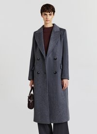 Cappotto doppiopetto in lana grigia scura. Presenta un colletto a risvolto, due tasche frontali e grandi bottoni neri. Il modello tiene in mano una borsa piccola.