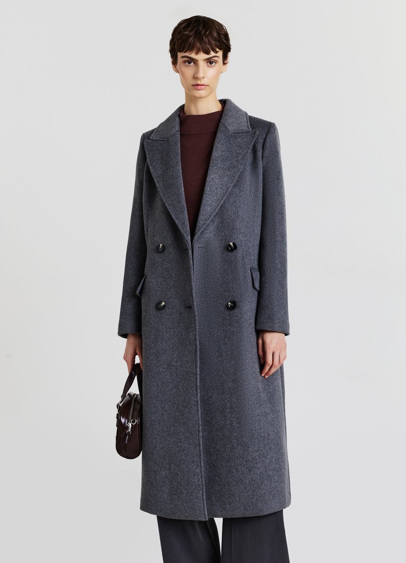 Cappotto doppiopetto in lana grigia scura. Presenta un colletto a risvolto, due tasche frontali e grandi bottoni neri. Il modello tiene in mano una borsa piccola.