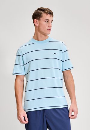 Fiatal férfi világoskék, vékony, vízszintes tengerészkék csíkokkal díszített rövid ujjú ingben és tengerészkék rövidnadrágban, jobbra nézve.