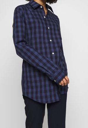 Osoba ubrana w granatową i czarną koszulę na guziki w kratę z długimi rękawami oraz ciemne spodnie, stojąca na jednolitym tle.