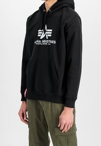 Felpa nera con cappuccio in misto cotone, dotata di tasca frontale, cappuccio con coulisse e logo bianco stampato con ali. Abbinata a pantaloni verde oliva.