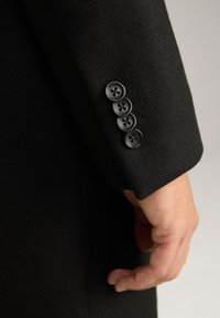 Manica di un blazer di lana nera con quattro bottoni rotondi neri con accenti floreali, che mette in risalto un tessuto texturizzato e una finitura sartoriale.