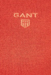 Pletený svetr v korálové barvě s vyšitým logem GANT v světle béžové barvě. Má jemné švy a měkkou, strukturovanou látku.