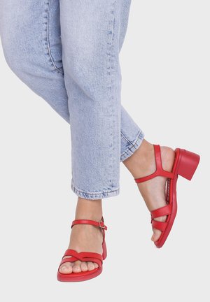Pies con sandalias rojas de tacón con tiras en los tobillos, cruzados en los tobillos, y vaqueros de mezclilla azul claro cortados por encima de los tobillos.