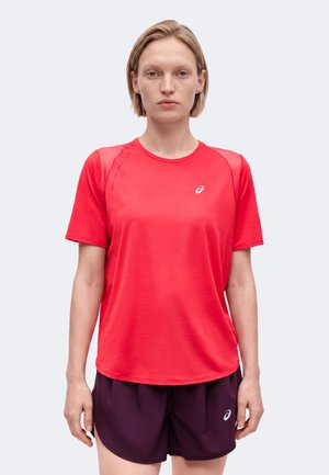 Femme aux cheveux blonds courts portant un t-shirt de sport rouge vif à manches courtes et un short de sport violet foncé, debout devant un fond uni.