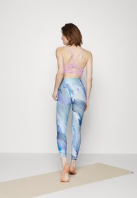 Bra de desporto rosa claro com alças e leggings azuis com padrão floral; modelo descalço, de costas, contra um fundo neutro.