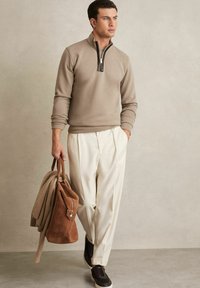 Beige vetoketjullinen pullover mustalla vetoketjulla, vaaleat housut, tummat lenkkitossut, pitää ruskeaa pukukangasta olevaa matkakassia ja villapaitaa. Sileä kangaspinta.