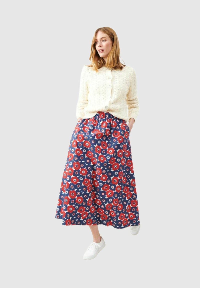 Kvinde iført en cremefarvet kabelstrikket cardigan, marineblå bukser med vid ben og rød blomsterprint, hvide sneakers, stående med hænderne i lommerne.