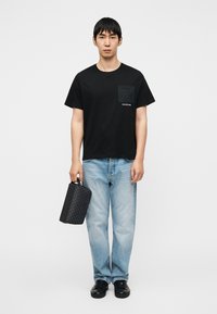 Μαύρο βαμβακερό μπλουζάκι με σχέδιο στην τσέπη στο στήθος, ανοιχτό μπλε τζιν παντελόνι και μαύρη τσάντα με σχέδιο και χειρολαβή, όλα σε λευκό φόντο.