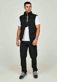 Jonge man staat opgesteld in een zwart gewatteerd vest, witte t-shirt, zwarte cargo broek en zwarte sneakers met witte veters.