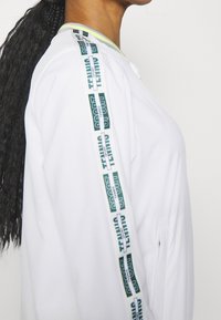 Vit sportjacka med siddragkedja, med grönt och svart logoband längs axelsömmen och en kontrasterande ribbad krage.